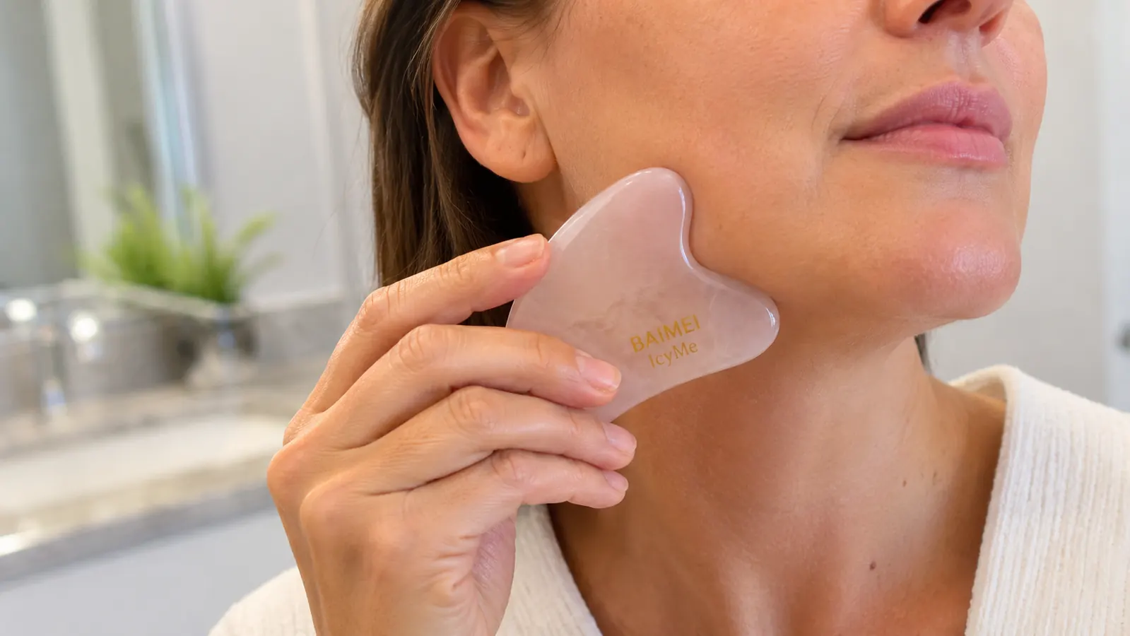 Close up of a hand using the BAIMEI rose quartz gua sha stone along the jawline