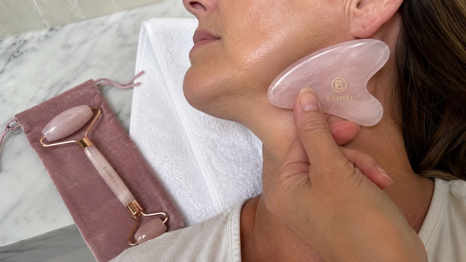 Close up of a hand using the BAIMEI rose quartz roller and gua sha stone on a jawline over a folded towel