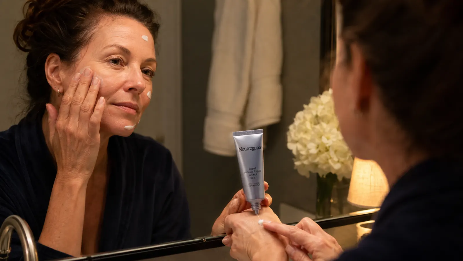 Hand applying retinol cream to the back of the hand near a bathroom mirror at night
